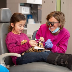 Young girl in a pink top sitting next to Dr. Pringle (Parr) holding a tiger toy and practicing flossing on it