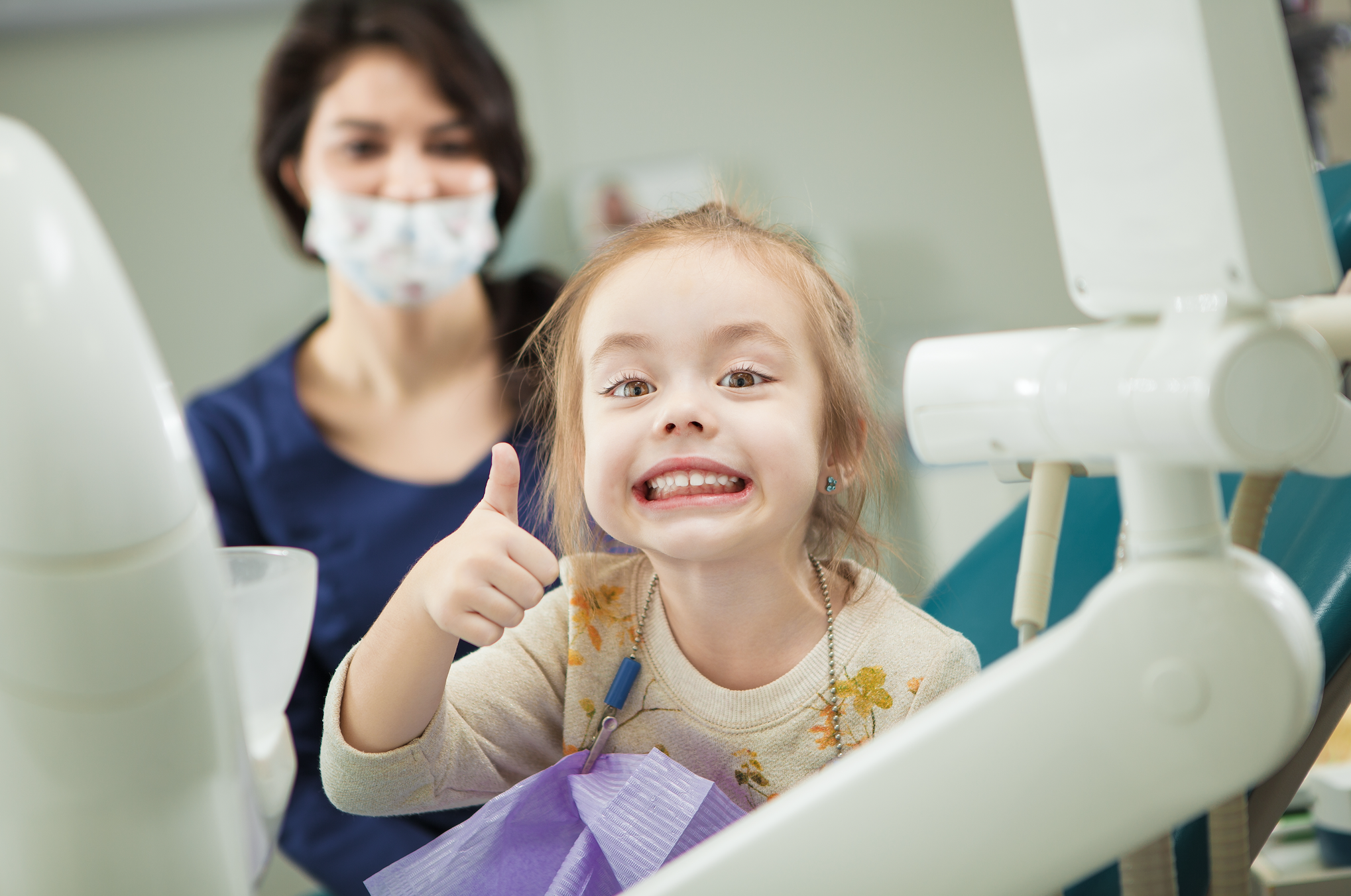 A young child sits in the dental chair, smiling with her thumbs-up sign.